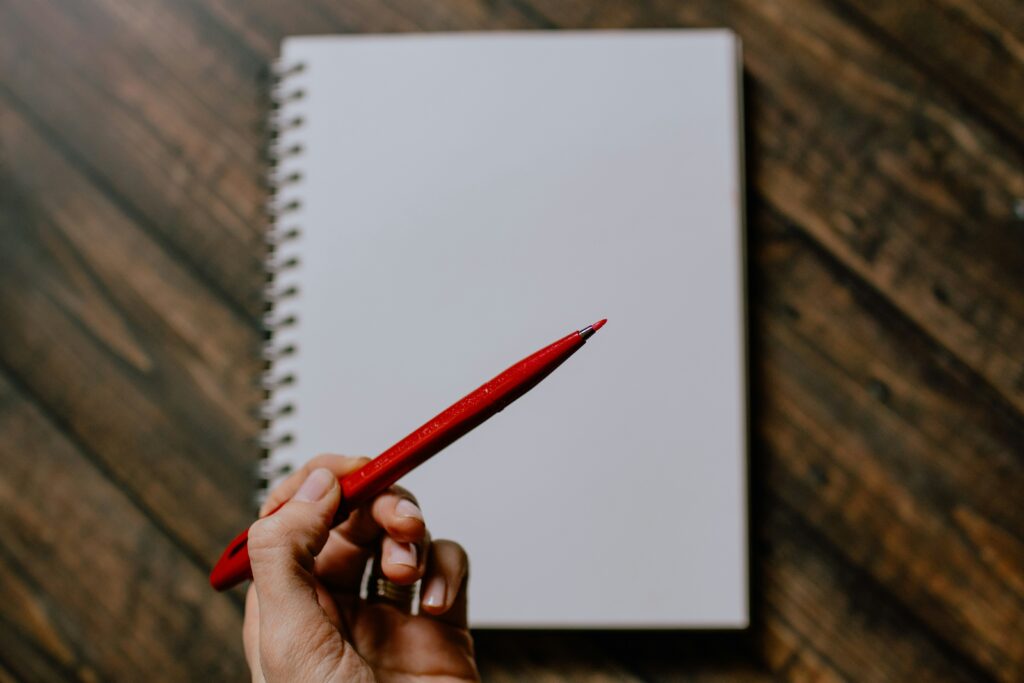 Hand holding a red pen over a blank spiral notebook on a wooden desk, representing editing and proofreading services